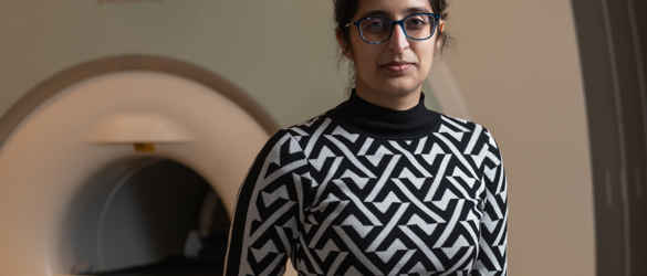 Dr Jasleen Jolly, stood in front of an MRI machine. She is wearing a black and white diamond pattern jumper and black glasses. She has dark hair and is holding the machine controls.