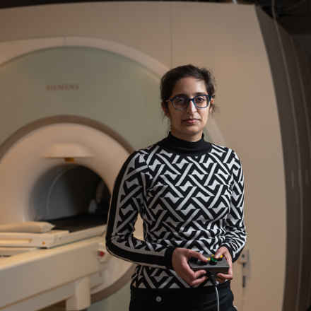 Dr Jasleen Jolly, stood in front of an MRI machine. She is wearing a black and white diamond pattern jumper and black glasses. She has dark hair and is holding the machine controls.