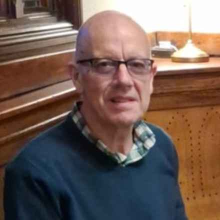 Macular Society volunteer Chris Norris, sat down wearing glasses and a shirt underneath a dark blue jumper.
