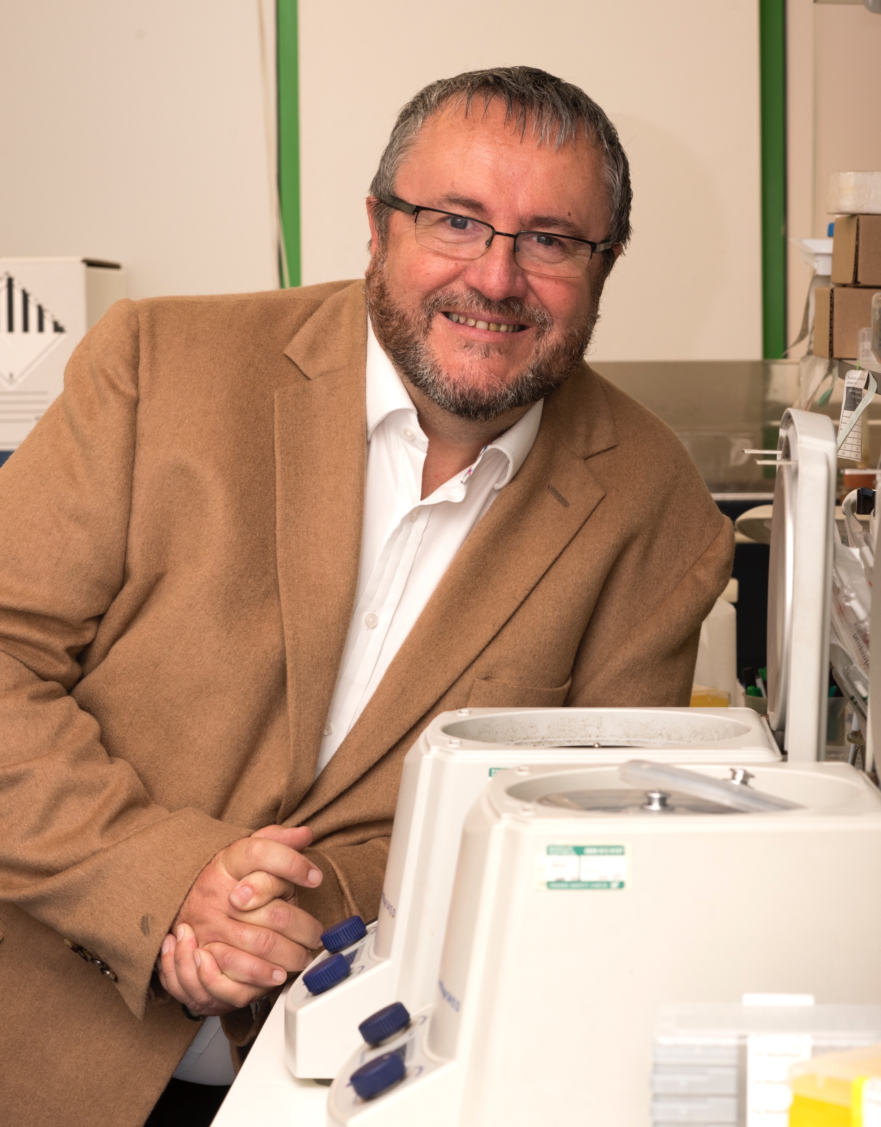 Pete Coffey, a white man in his 60s with grey hair and a beard wearing a brown suit jacket smiling and leaning against lab equipment
