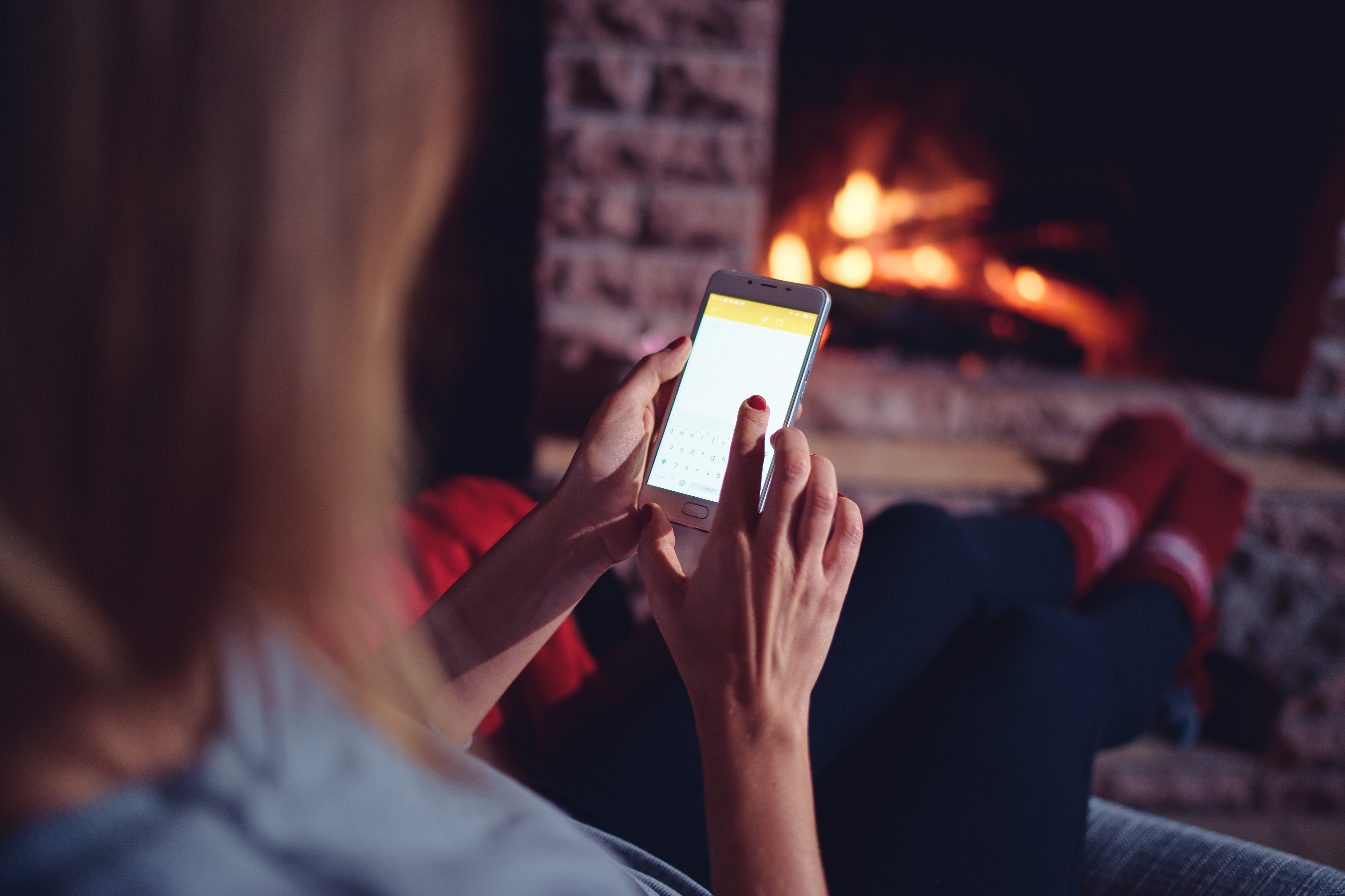 Winter Warmer. Woman on smart phone by cozy fire.