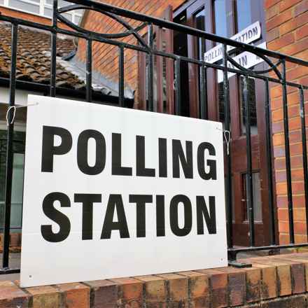 Polling station sign hanging on a black railing with a brick building in the background
