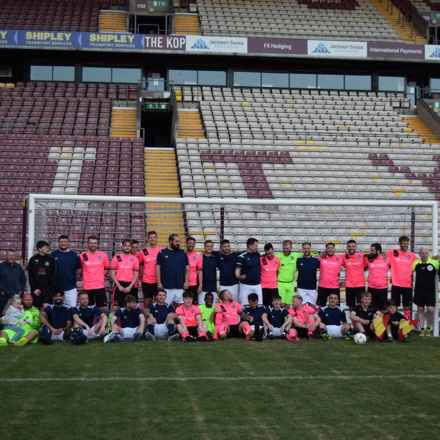 Football players of Craven For Change FC fill the goal in front of the stand at Bradford's Valley Parade Stadium, as they get together for a charity match in aid of the Macular Society. Players are wearing either kits of black shirts and white shorts, or pink shirts and black shorts. Goalkeepers are wearing all-green kits.